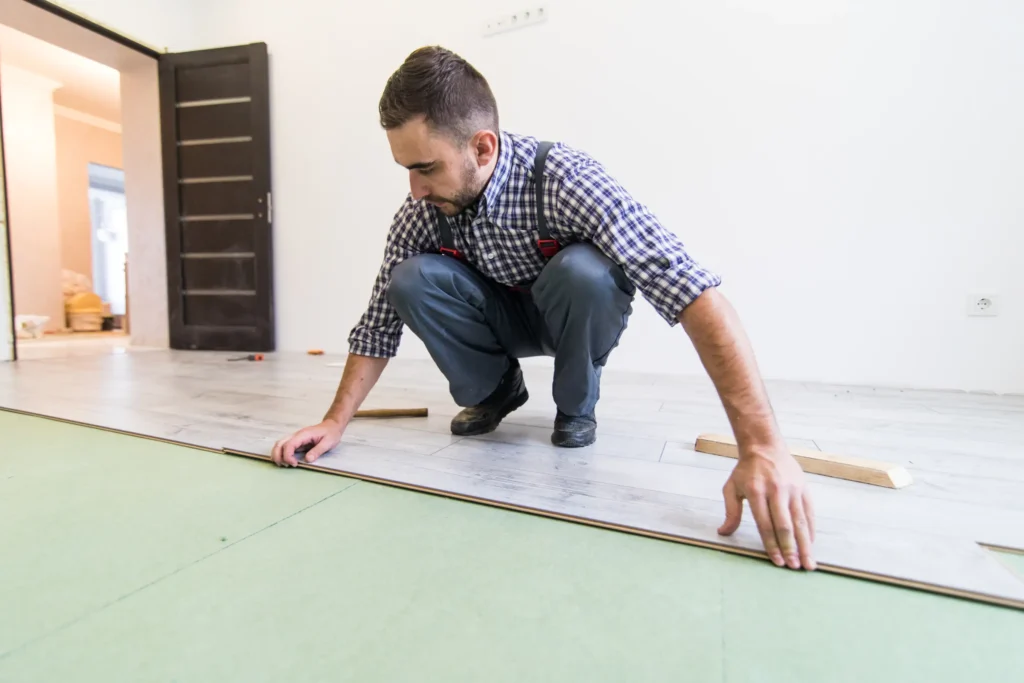 Close-up overhead view of a worker kneeling and laying light-colored laminated flooring boards over a green underlayment