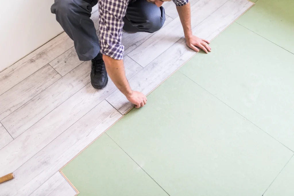 A worker, kneeling, is installing a long plank of light-gray laminated flooring