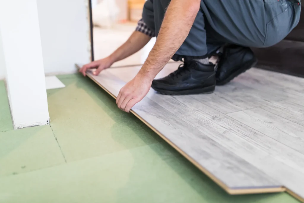 A young male worker in a plaid shirt and dark trousers kneels on a partially installed light laminate floor